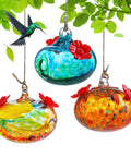 Set Of 3 Small Blown Glass Hummingbird Feeders For Outdoors Hanging With 2 Flower Feeding Ports 3 Inch Glass Hummingbird Feeders Garden Decor For Outside, multicolored with red flower ports, hanging among leaves.