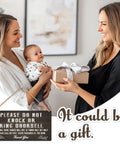 Two women exchanging a gift with a baby and the Please Do Not Ring Baby Sleeping Sign for Front Door hanging nearby.