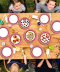 48 Pack Red And White Checkered Disposable Paper Plates For Backyard Kickbacks arranged on a wooden table with food and drinks.
