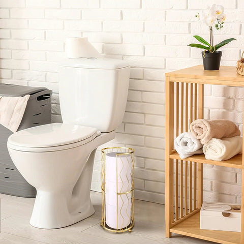 Gold Finish Standing Toilet Paper Holder With Three Roll Reserve next to a toilet in a bathroom with wooden shelf and towels