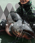 Person holding large catfish in water, wearing camouflage jacket, showcasing catch from Six Piece Catfish Drag Weights Sinker Kit No Snag Design.