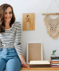 Woman sitting on a bench next to books and a macrame wall hanging with a wooden light fixture mounted on the wall.