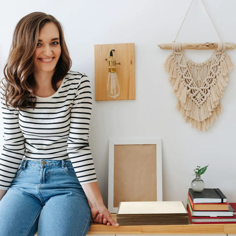 Woman sitting on a bench next to books and a macrame wall hanging with a wooden light fixture mounted on the wall.