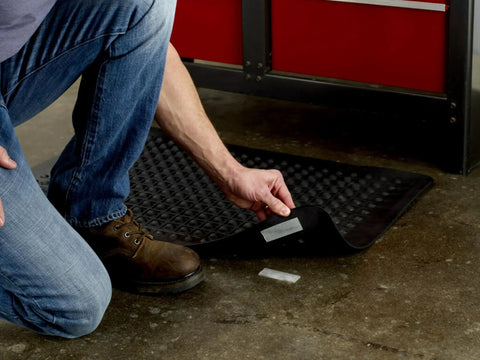 Person installing heavy duty reclosable fastener roll with weatherproof adhesive under a black mat on a concrete floor.