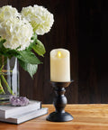 Realistic Moving Flame LED Candle Pillar With Scalloped Edge And Smooth Finish For A Cozy Glow Vanilla Honey Scented Ivory Timer Function Battery Operated 3 Inch By 4.5 Inch on black stand beside flowers on wooden table