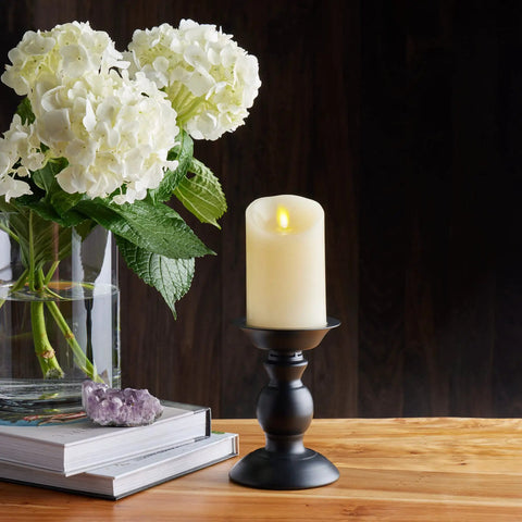 Realistic Moving Flame LED Candle Pillar With Scalloped Edge And Smooth Finish For A Cozy Glow Vanilla Honey Scented Ivory Timer Function Battery Operated 3 Inch By 4.5 Inch on black stand beside flowers on wooden table