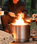 People cooking with Portable Pizza Oven Firewood Logs for Outdoor Cooking 6 Inch White Oak Kiln Dried Low Smoke Fuel for Ooni Karu 12 Solo Stove Pi and Bertello. Logs stacked beside campfire.