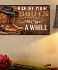 Decorative sign reading 'Kick Off Your Boots and Stay A While' with cowboy boots and hat next to a lamp and flowers.