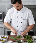 Unisex Chef Coat With Short Or Long Sleeve Crafted From Breathable Cotton Poly Blend worn by chef chopping herbs at kitchen prep station.