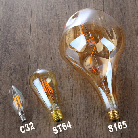 Oversized Vintage LED Edison Bulb With 2200K Warm White Glow Dimmable 5W 400LM Amber Glass Giant Irregular Shape For Pendant Or Floor Lamps E26 Base Decorative Extra Large Bulb For Cozy Retro Lighting next to smaller bulbs on wooden surface.