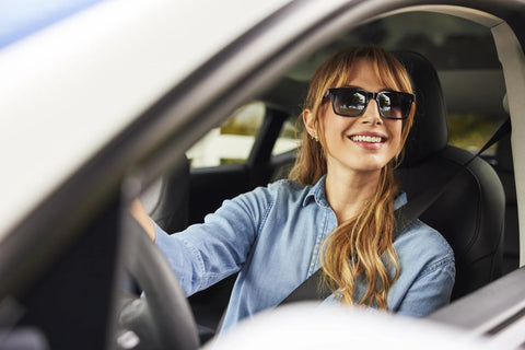 Sunglasses on smiling woman driving a car; black frames, denim shirt, seat belt