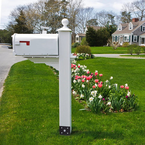 White mailbox with red flag on decorative post near garden with blooming tulips in residential neighborhood setting.