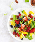 Fresh salad with strawberries, blueberries, raspberries, mango, pecans, and greens on a white plate with pecans on the side.