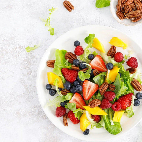 Fresh salad with strawberries, blueberries, raspberries, mango, pecans, and greens on a white plate with pecans on the side.