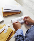 Person applying Hassle Free Direct Thermal Mailing Address Labels In Bright White, Sized 1-1/8 X 3-1/2 Inches to an envelope.