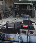 2000 Lb 12V Electric Winch With Steel Cable, Hook And Corded Remote Portable Compact Power in a workshop with chains and vehicle