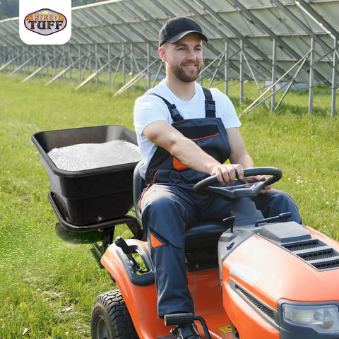 Man operating utility tractor with 12V Grass Seed And Fertilizer Spreader With 80 Pound Capacity Hitch Mount Receiver in field.