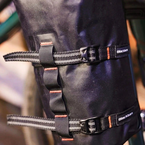 Close-up of a black Outpost Cargo Water Bottle Cage with triple mount holes and straps securing gear on a bike frame.