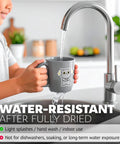 Person holding a mug with a cat sticker under running water, demonstrating water-resistant vinyl sticker paper use.