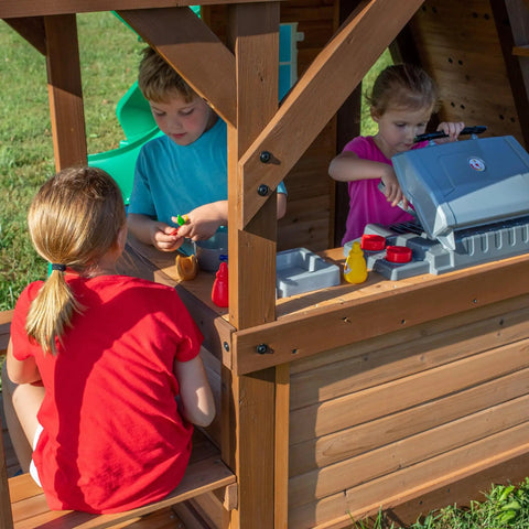 Children playing in Cedar Cove Wooden Swing Set With Spiral Tube Slide And Wave Slide, Elevated Clubhouse With Balcony, Toy Grill