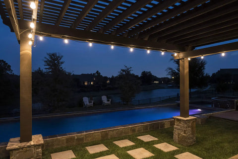 Backyard Bistro Style 24 Foot Outdoor String Lights illuminating a pergola by a pool at night, 12 shatter-resistant bulbs visible