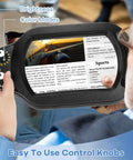 Person using a 10X Rectangle Magnifying Glass With Light And Stand For Close Work And Reading Hands Free LED Desk Lamp With Clamp Real Glass Lens 10X Magnification 5 Color Modes Stepless Dimmable 2 In 1 Lighted Magnifier Desk Lamp to read a newspaper, highlighting its brightness and color modes with control knobs visible.
