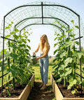 Woman watering plants under Metal Core Garden Arch Trellis For Climbing Plants Outdoor 87 Inch Tall 71 Inch Long 75 Inch Wide Archway Tunnel Plant Support For Vines Cucumbers Tomatoes Grapes And Vegetables Durable Space Saving Ground Or Raised Bed Friendly.