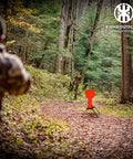 Shooter aiming at AR500 Steel Auto Reset Target Thirteen Inch Popper in forest setting, self-reset gong for practice.