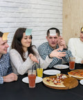 Group of friends playing a game with sticky notes on foreheads, sitting around a table with pizza drinks in a casual indoor setting.