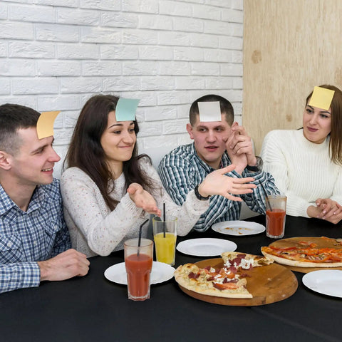 Group of friends playing a game with sticky notes on foreheads, sitting around a table with pizza drinks in a casual indoor setting.