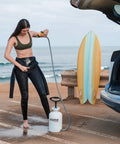 Woman using High Pressure 5L Portable Shower Kit Hand Pump 45 PSI No Power Needed for Camping next to car and surfboard at beach