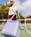 Woman carrying Lilac Breeze 11 Qt Tag-Along Cooler With Strapped Crossbody Carry, Leakproof Lid Locks, MaxCold Insulation on tennis court.