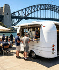 Ready To Roll 13 Foot Vintage Food Trailer With Fully Equipped Mobile Catering Kitchen And Slick White Exterior For Serious Commercial Food Truck Hustles, parked by a bridge with customers.