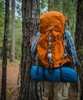 Person hiking in forest with orange backpack, blue sleeping bag attached, wearing shorts and plaid shirt, trees surrounding.