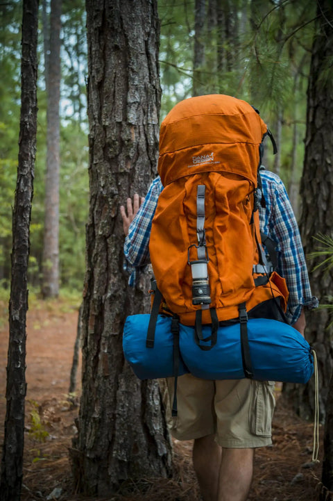 Person hiking in forest with orange backpack, blue sleeping bag attached, wearing shorts and plaid shirt, trees surrounding.