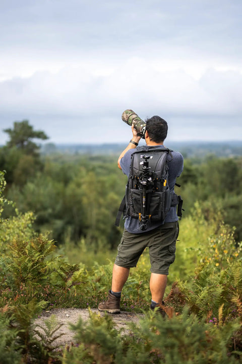 Photographer using Alta Sky 62 Camera Backpack For 200 To 600mm Telephoto On DSLR Or Full Frame Mirrorless Fits Up To 7 Lenses Plus Beefed Up Gear 16 Inch Laptop And 11 Inch Tablet Top Front And Lower Rear Access Room For Tripod in nature setting.