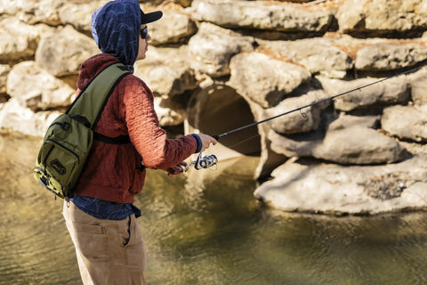 Angler wearing Mystery Tackle Box Pond Prowler Sling Fishing Lure Kit Green Sling Pack fishing near rocks.