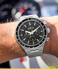 Run The Street With This Badass 44 Mm Men's Watch Featuring A Gearshift Inspired Face Premium Stainless Steel Bracelet And Bold Black Dial For Daily Quartz Performance on a wrist next to a car interior.