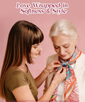 Young woman tying floral scarf on older woman against pink background