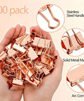 Hands holding rose gold binder clips, 100 pack, featuring stainless steel handles and solid metal material with arc corners.