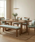 Dining room with wooden table, chairs, and a bench featuring floral cushions. Sunlit window in background.