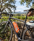 Easy Squeeze Cycling Water Bottle 680 ML Lightweight Bike Hydration Flask in orange on bicycle by a stone wall.