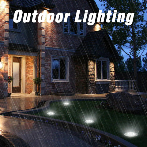 12 Pack Solar Ground Lights With 8 LED Each In Ground Round Puck Lights illuminate a rainy pathway by a house at night.