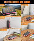 Outdoor Power Bank 33800mAh With Solar Charging And Super Bright LED Flashlight Portable Charger With Built In Cables Wireless Charging And Compass 7 Outputs 4 Inputs For Camping And Hiking showcasing multiple charging options including solar, USB-C, USB A, and micro USB on a desk and outdoor settings.