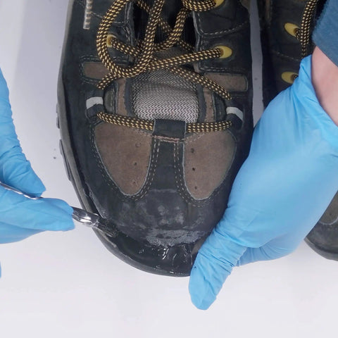 Applying Aquaseal SR Shoe Repair Adhesive to the sole of a black hiking boot with blue gloved hands demonstrating the repair process.