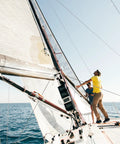 Two people adjusting sails on a sailing boat in open water, clear sky overhead, side view of the boat and sails.