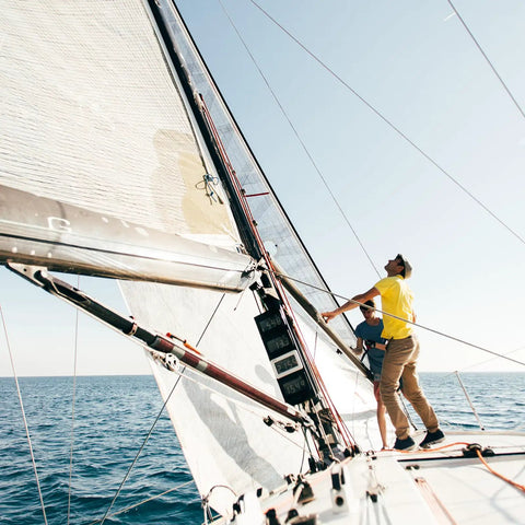 Two people adjusting sails on a sailing boat in open water, clear sky overhead, side view of the boat and sails.