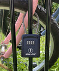 Rugged Outdoor Key Lock Box With Cable Shackle, Small Waterproof Metal Safe, 4 Digit Combo For Spare Keys on fence with bike