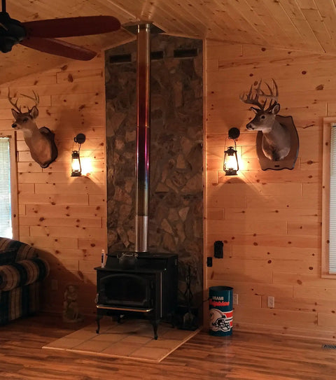 Rustic Wall Sconce Lantern With Glass Globe For Farmhouse Decor And Old School Vibe LED Five Point Five Watt Candle Light Electric Wall Light Fixture In Rust Finish With Authentic Oil Lantern Reproduction Style And Bottom Lit Glow next to fireplace with deer mounts in cabin setting