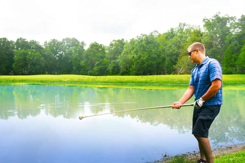 Man using Compact Stainless Steel Golf Ball Retriever With 10-Foot Length And 14-Foot Reach near a pond on a golf course.